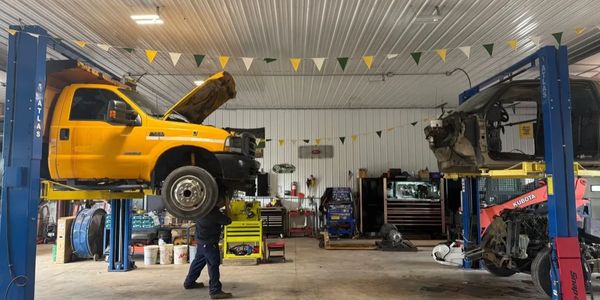 Two trucks lifted on hoists in a spacious garage with a mechanic working.