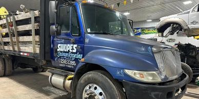 Blue salvage recycling truck parked inside a garage with a dismantled vehicle nearby.