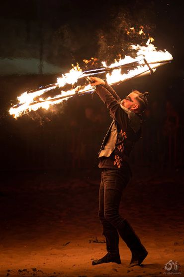 A man performing a fire dance with flaming props at night on sandy ground.