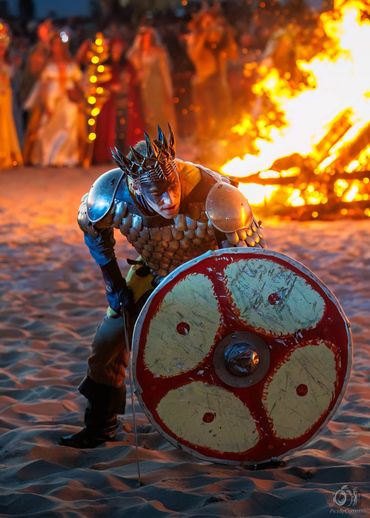A warrior in armor with a large shield poses near a bonfire on sand.