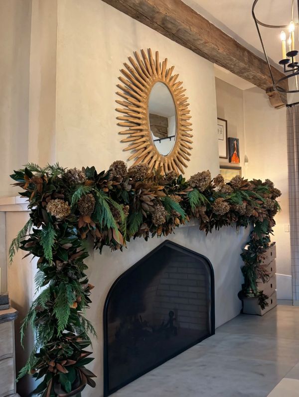 Cozy fireplace with a rustic sunburst mirror and lush green garland.