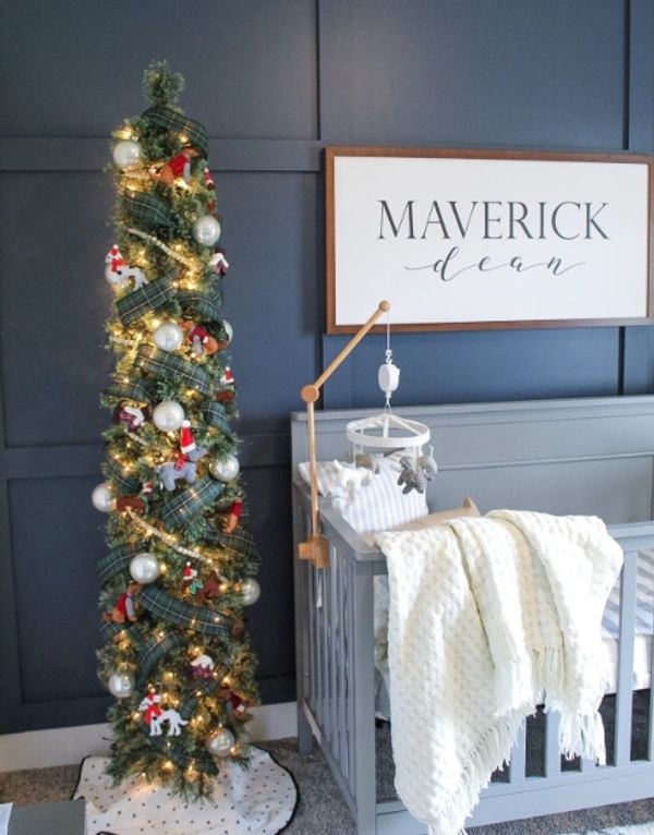 Slim Christmas tree with lights and ornaments beside a gray baby crib.