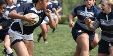 Women playing rugby, one holding the ball while others chase her.