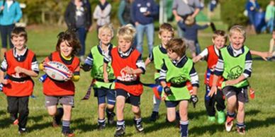 Children playing rugby in colorful vests outdoors on a sunny day.