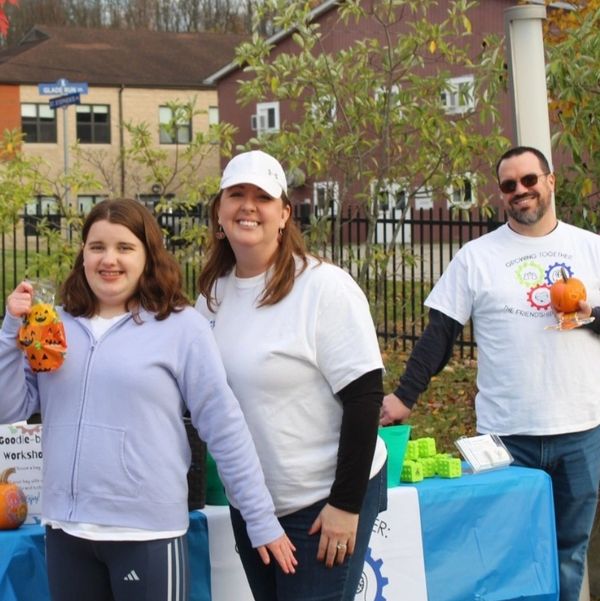 Three people smiling outdoors holding Halloween-themed items near a blue table.