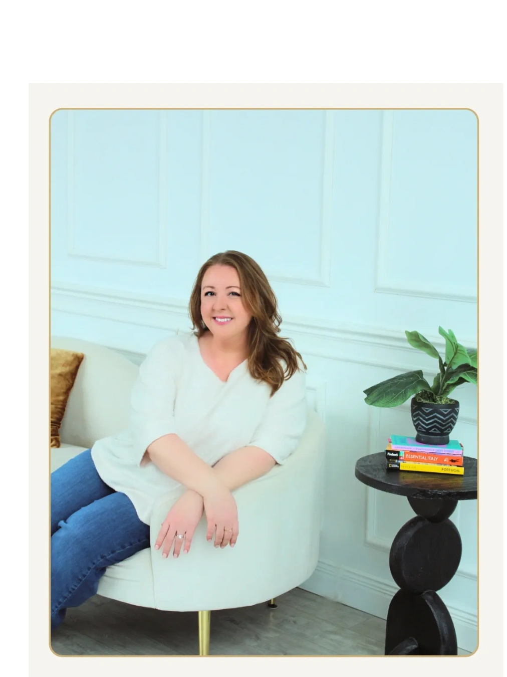 Woman sitting on a small couch with arms crossed at wrists, smiling next to table with travel books.