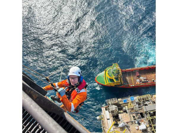 Worker in safety gear rappelling down an offshore platform with a supply ship nearby.