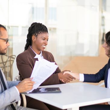 Two women shaking hands in a business meeting while a man reviews documents.