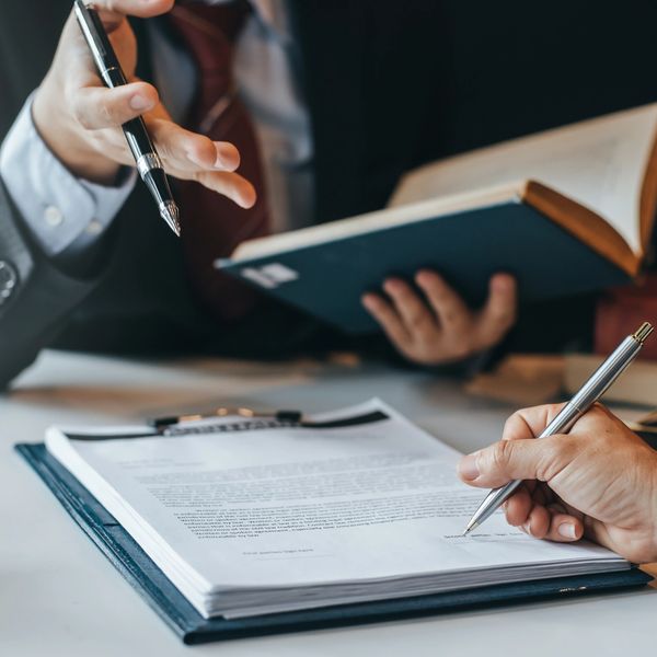 Person signing a document while another explains with a book.