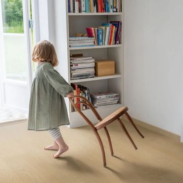 A child carries a wooden chair backwards in a cozy room with a bookshelf.