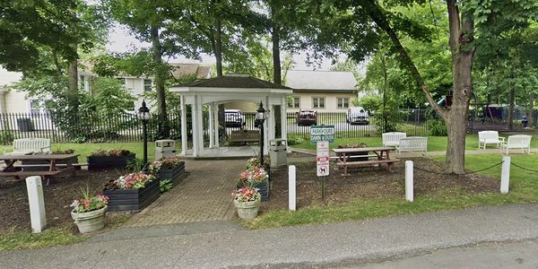 Walkway leads ups to a small white gazebo surrounded by potted plants and blenches