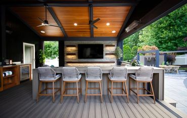Modern outdoor bar area with five wicker stools and a mounted TV under a wooden ceiling.