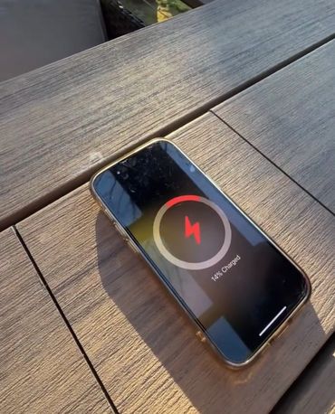 Smartphone charging with 14% battery on a wooden surface in sunlight.