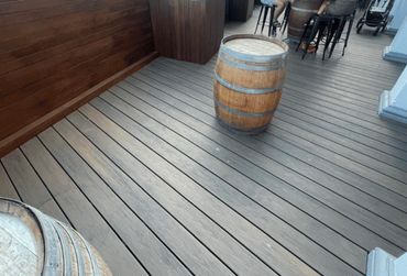 Wooden barrels on a wooden deck with people sitting on stools in the background.