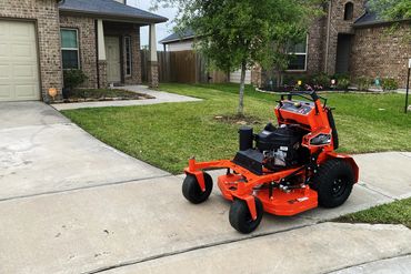 A bright orange zero-turn lawn mower parked on a driveway near a green lawn.