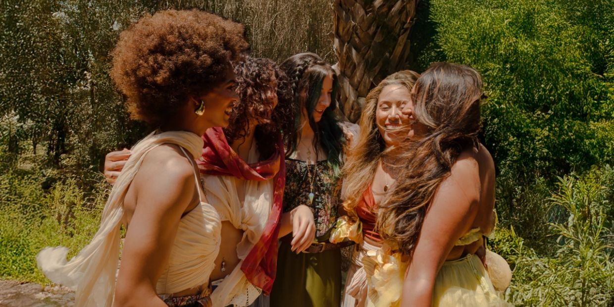 A group of five women joyfully embracing outdoors in sunny nature.
