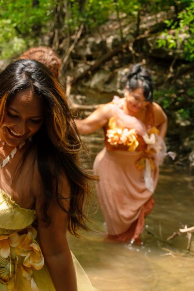 Two women in floral dresses wading through a forest stream.