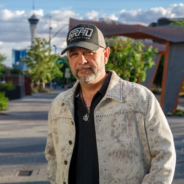 Man in a stylish jacket and cap standing outdoors on a sunny day.