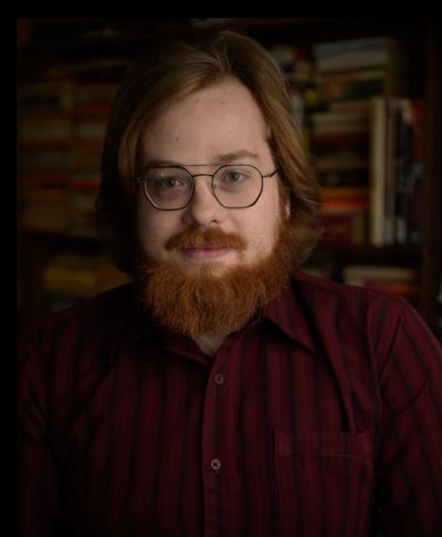 Bearded man with glasses and a red shirt, smiling gently indoors.