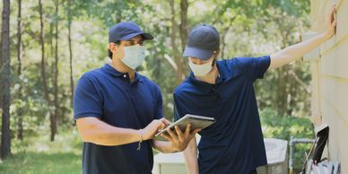 Two masked technicians in blue uniforms inspecting an air conditioning unit outdoors.
