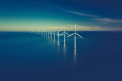 A line of offshore wind turbines stretches across the ocean under a clear sky.