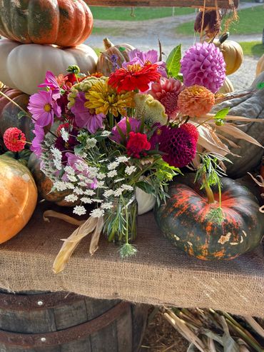 Colorful autumn flowers in a vase surrounded by pumpkins on a burlap-covered table.