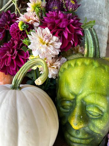 A green pumpkin shaped like a human face next to a white pumpkin and colorful flowers.