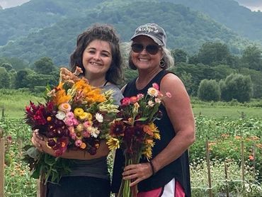Two women smiling while holding colorful flower bouquets in a lush garden.