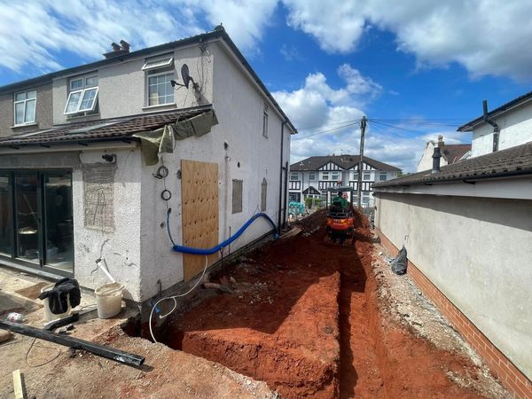 Construction work digging foundation beside a house under a partly cloudy sky.