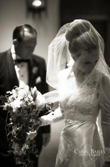 A bride in a lace wedding dress holding a cascading bouquet, with a man in a tuxedo behind her.