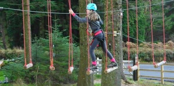 Girl crossing rope bridge with safety gear in forest.