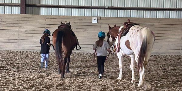 Two children walking horses in an indoor arena.