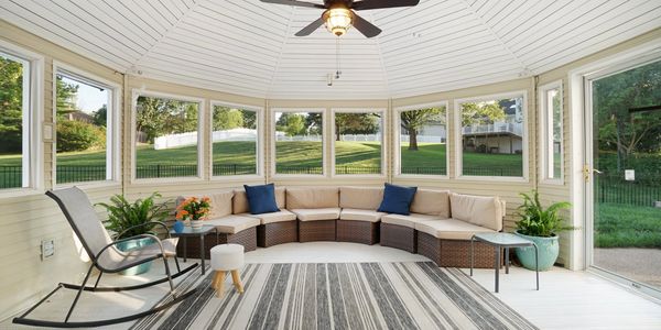 Sunroom with neutral sectional, rocking chair, live plants, striped neutral rug, and ceiling fan.
