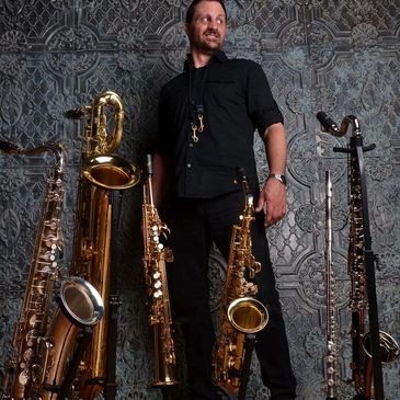 Man stands confidently among various saxophones against a textured wall.