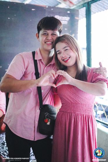A smiling couple in pink making a heart shape with their hands indoors.