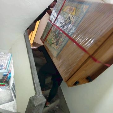 Two people carrying a wrapped wooden cabinet upstairs in a narrow stairway.