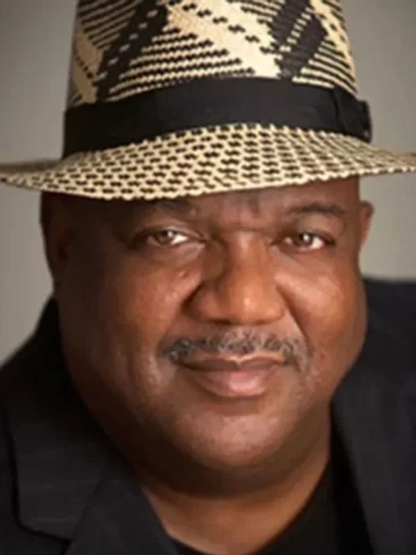 Smiling man wearing a patterned fedora hat and black jacket.