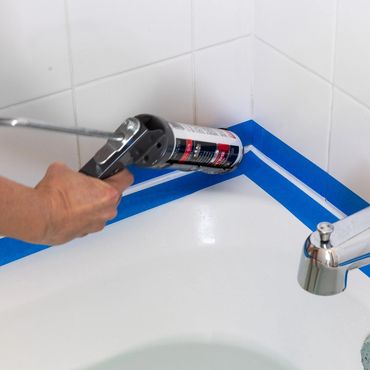 Applying caulking around a bathtub with blue painter's tape for clean edges.