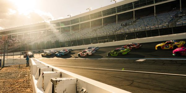 Race cars speeding on a sunlit racetrack with grandstands in the background.