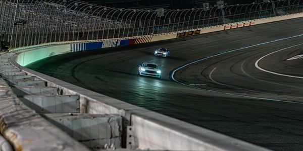 Two cars race on a curved track at night under bright headlights.