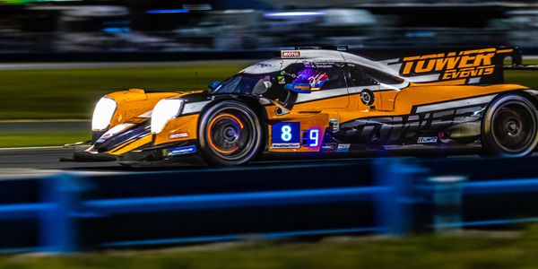 Orange and black race car speeding on a track at night with headlights on.