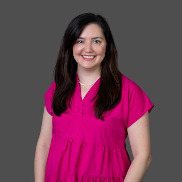 Smiling woman in a bright pink blouse against a gray background.