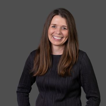 Smiling woman with long brown hair wearing a dark ribbed sweater against a gray background.
