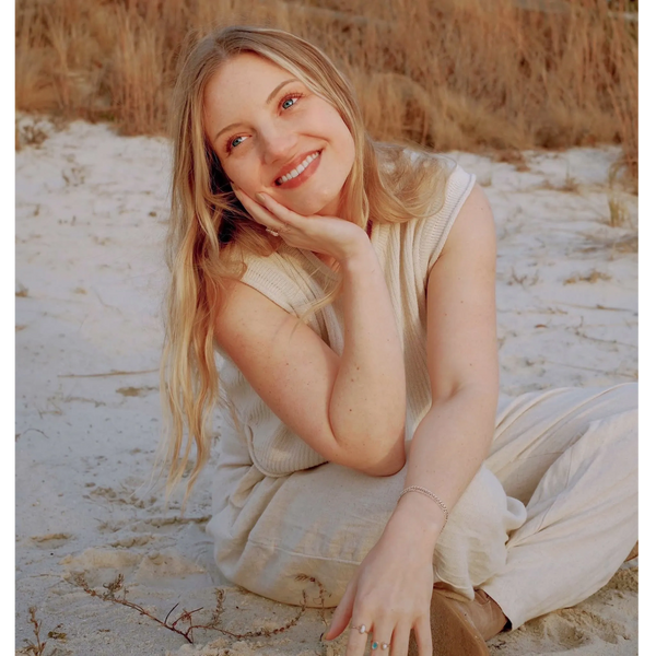 Smiling woman with blonde hair sitting on sandy ground in a cozy beige outfit.