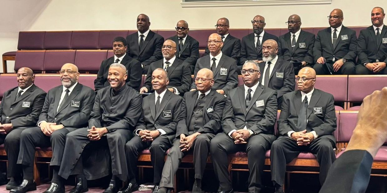 Group of men in black suits seated in rows inside a church or hall.