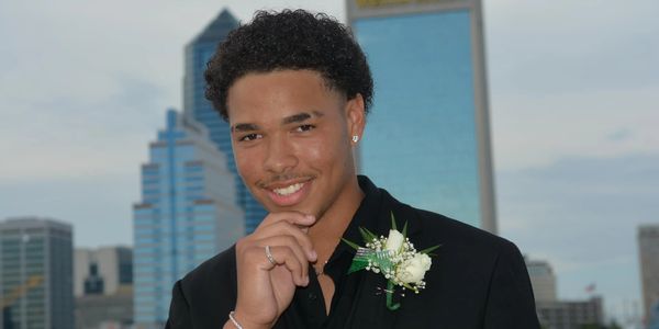 Young man in formal attire with a boutonniere smiles against a city backdrop.