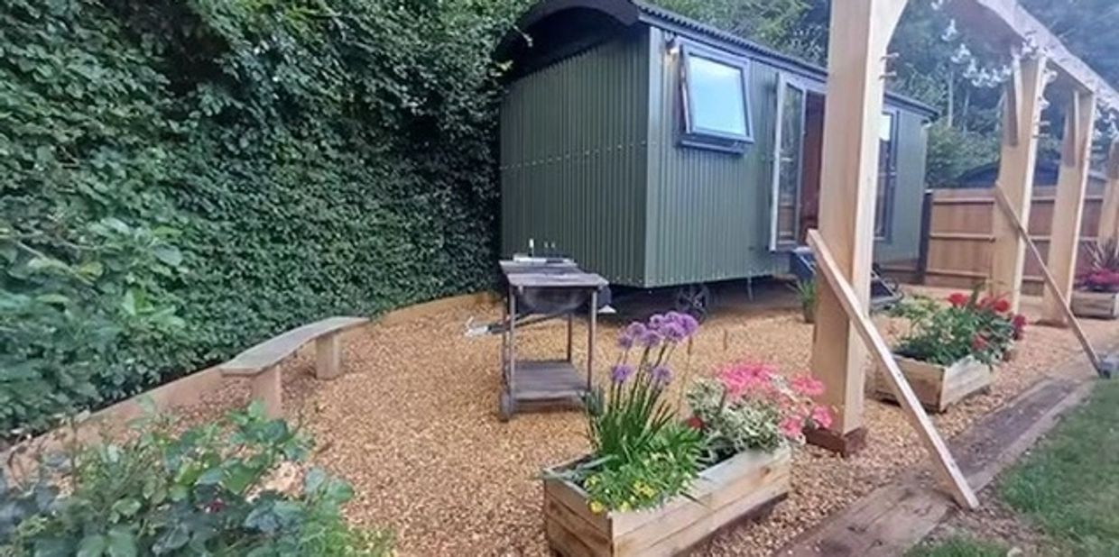 A small green cabin with outdoor plants and a gravel area.