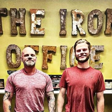Two men in red shirts standing in front of a rustic sign.