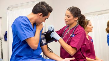 A nurse in maroon scrubs checks a patient's blood pressure in a clinical setting.