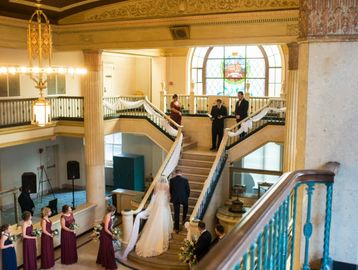 Bride and groom walk up grand staircase with wedding party in elegant venue.
